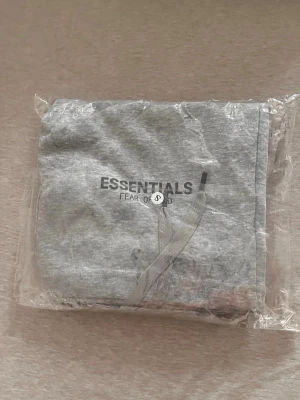 Grå Essentials Fear of God shorts - Säljer ett par grå shorts från Essentials Fear of God med snörning i midjan och två sidofickor. Klassisk logga tryckt på vänster ben. Sköna och stilrena shorts i mjuk bomullsblandning, perfekta för sommaren.