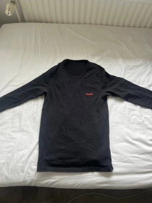 Svart långärmad topp från Hugo - Svart långärmad topp från Hugo med rund halsringning och diskret röd logga på bröstet. Tillverkad i mjuk bomull som känns skön mot huden. Enkel och stilren design som passar till det mesta.