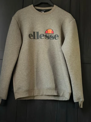 Grå sweatshirt från Ellesse - Snygg grå sweatshirt från Ellesse med klassisk logga i svart, orange och rött framtill. Tröjan har rund halsringning, långa ärmar och ribbade muddar vid ärmslut och nederkant. Perfekt för en avslappnad och sportig stil.
