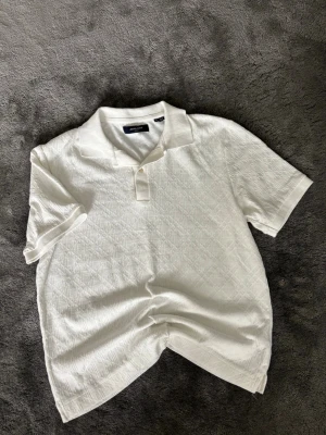 Vit piké från Jack & Jones - Säljer en stilren vit pikétröja från Jack & Jones med diskret strukturmönster. Tröjan har klassisk krage, korta ärmar och knappar vid halsen. Perfekt till jeans eller shorts för en clean look.