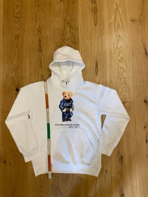 Vit hoodie Polo Bear Ralph Lauren - Vit hoodie från Polo Bear by Ralph Lauren med ikoniskt björnmotiv på bröstet. Klassisk känguruficka framtill, ribbade muddar och huva med dragsko. Perfekt för dig som gillar streetwear och vill ha en chill look.