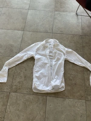 Vit klassisk skjorta med lång ärm - Säljer en stilren vit skjorta med lång ärm och klassisk krage. Skjortan har knappar framtill och är tillverkad i ett lätt bomullstyg. Perfekt för dig som gillar en clean och enkel look.