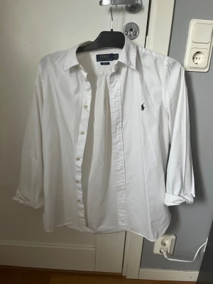 Vit skjorta från Polo Ralph Lauren - Klassisk vit skjorta från Polo Ralph Lauren med den ikoniska broderade loggan på bröstet. Skjortan har lång ärm, knappar framtill och är gjord i mjuk bomull. Perfekt till jeans eller chinos för en clean och stilren look.
