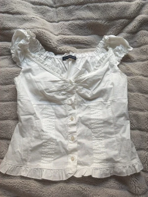 Vit blus med volang från Brandy Melville - Söt vit blus från Brandy Melville med knappar framtill, volangdetaljer vid ärmslut och nederkant samt lätt rynk vid bysten. Blusen har korta puffärmar och en romantisk känsla. Perfekt till jeans eller kjol.