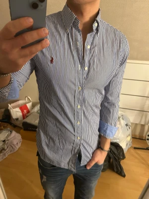 Randig skjorta från Ralph Lauren - Säljer en klassisk blåvit randig skjorta från Ralph Lauren med button-down krage och broderad logga på bröstet. Skjortan har långa ärmar, vita knappar och kontrasttyg i ljusblått på insidan av manschetterna. Perfekt för en stilren look.