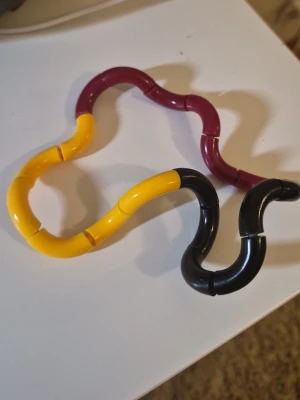 Fidgetleksak i plast, gul svart vinröd - En böjbar fidgetleksak i plast med länkar i färgerna gul, svart och vinröd. Perfekt att snurra och forma i olika mönster när man vill ha något att pilla med. Lätt att ta med i fickan eller väskan.