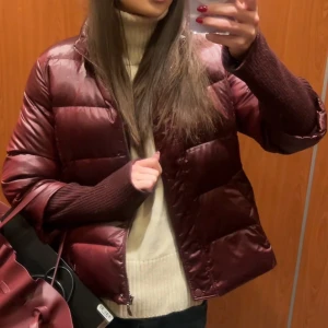 Vinröd pufferjacka med ribbade muddar - Snygg vinröd pufferjacka med breda, quiltade paneler och långa ribbade muddar i matchande färg. Jackan har hög krage och dragkedja framtill. Perfekt för våren. Sitter som en M/S