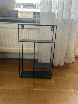 Svart metallhylla med tre plan - Snygg svart hylla i metall med tre olika hyllplan i asymmetrisk design. Perfekt för småprylar, dekorationer eller växter. Hyllan har två upphängningshål upptill för enkel montering på väggen. Stilren och modern look som passar i de flesta rum.