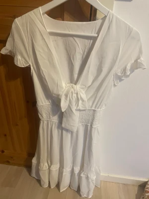 Vit klänning  - Söt vit kortklänning med omlottdesign och knyt framtill. Klänningen har volangdetaljer i kjolen och korta ärmar. Perfekt för sommarens alla tillfällen och ger en luftig och fräsch känsla. Tillverkad i ett lätt material som känns bekvämt mot huden.