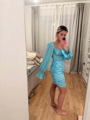 Adoore Blå miniklänning med puffärmar - Säljer en ljusblå miniklänning med långa puffärmar och volang vid ärmslut. Klänningen har rynkade detaljer längs sidorna. Fyrkantig urringning och snörning i sidan. Knäpps med dragkedja på sidan. 