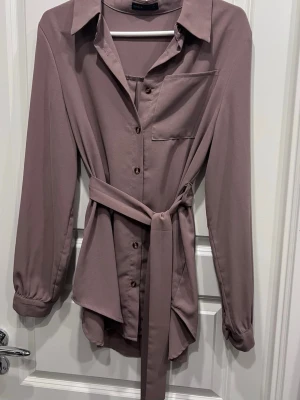 Ljusbrun/lila skjorta med bälte från SHEIN  - Stilren ljusbrun/lila skjorta från SHEIN med lång ärm, klassisk krage och knappar framtill. Skjortan har ett matchande bälte i midjan och en bröstficka. Perfekt för en trendig och avslappnad look.