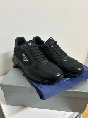 Svarta sneakers från Prada - Snygga svarta sneakers från Prada med diskret logga på sidan. Skorna har en stilren design i en mix av läder och textil, svarta skosnören och robust sula. Perfekta för dig som gillar exklusiva och minimalistiska sneakers.