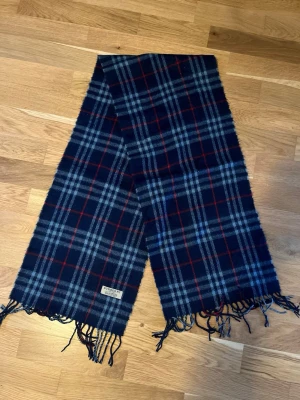 Blårutig halsduk från Burberry - Snygg halsduk från Burberry i blått med rutigt mönster i rött och vitt. Halsduken har fransar i ändarna och är tillverkad i mjuk ull. Perfekt accessoar för att lyfta din outfit med klassisk brittisk stil. Nypris ca 5000 kronor. Pris går att diskuteras vid snabb affär