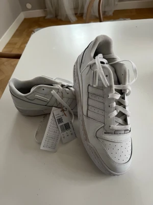 Vita Adidas sneakers med retrostil - Snygga vita sneakers från Adidas med klassiska tre ränder på sidan och perforerade detaljer framtill. Skorna har en låg profil, rund tå och robust yttersula med cirkulärt mönster. Tillverkade i läder och textil, perfekta för en clean och sportig look.