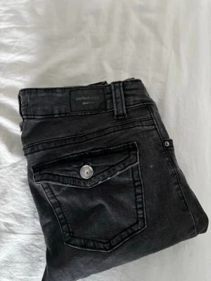 Svarta jeans från Gina Tricot - Säljer ett par svarta lågmidjade jeans från Gina Tricot. De har klassisk femficksdesign med fickor med knapp bak, bootcut ben och silverfärgade detaljer. Materialet är stretchigt denim som sitter snyggt och bekvämt.