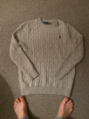 Beige kabelstickad tröja Polo Ralph Lauren - Kabelstickad beige tröja från Polo Ralph Lauren med rund halsringning och klassisk broderad logga på bröstet. Tröjan har långa ärmar och ribbade muddar vid ärmslut och nederkant. Perfekt för lager-på-lager och en stilren look. Det står M men den passar även S