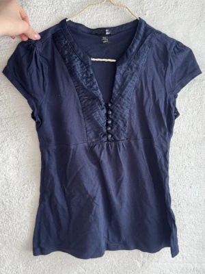 Mörkblå topp med knappar - Mörkblå topp från H&M med korta ärmar och dekorativa knappar framtill. Toppen har ett snyggt veckat parti vid halsringningen och är tillverkad i mjuk bomull. Perfekt till jeans eller kjol för en enkel men stilren look.