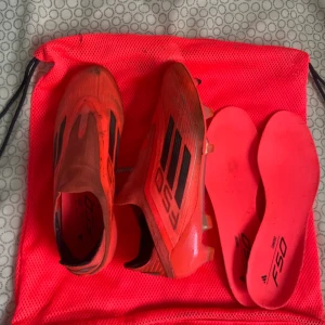 Adidas F50 röda fotbollsskor - Säljer ett par Adidas F50 fotbollsskor i rött med svarta detaljer och klassiska tre ränder på sidan. Skorna har en strömlinjeformad design och kommer med matchande röda innersulor. Perfekta för dig som vill ha lätta och snabba skor på planen.