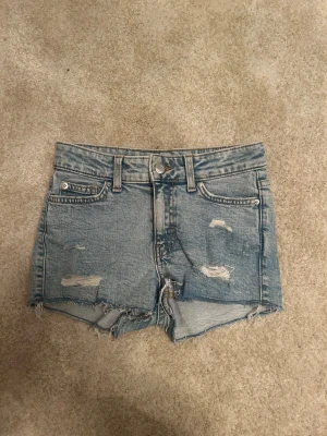 Blå slitna jeansshorts - Säljer ett par ljusblå jeansshorts med slitna detaljer och råa kanter. Klassisk femficksmodell med bälteshällor och knappgylf. Perfekta för varma dagar och en avslappnad look. Dem är i storlek 134 