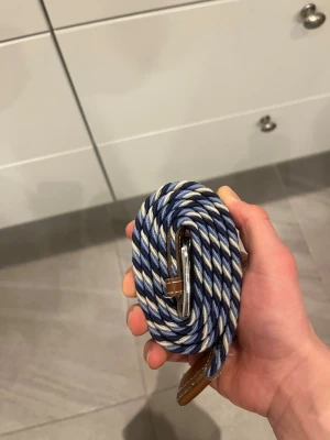 Flätat blåvitt bälte med läderdetaljer 105 cm - Säljer ett snyggt flätat bälte i blått, vitt och beige med bruna läderdetaljer och silvrigt spänne. Perfekt för att ge din outfit en cool touch. Bältet är elastiskt och passar till både jeans och chinos. 105 cm. Inga skador eller tecken på användning.