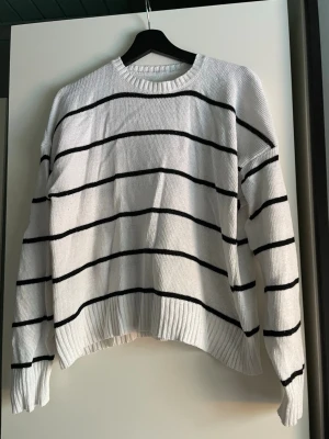 Vit stickad tröja med svarta ränder - Säljer en vit stickad tröja med tunna svarta horisontella ränder. Tröjan har rund halsringning, långa ärmar och ribbade muddar vid ärmslut och nederkant. Perfekt för en avslappnad och stilren look. Någon noppra finns. Innehåller lite ull, så den värmen något. Storleksetikett saknas men känns som M