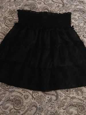 Svart volangkjol med smock - Supersöt svart kortkjol med bred resår i midjan och flera lager volanger. Kjolen har en lätt struktur i tyget och ger en fluffig look. Perfekt att styla med en enkel topp eller stickad tröja. Kan skicka fler bilder vid förfrågan 