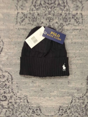 Svart mössa Polo Ralph Lauren - Snygg svart kabelstickad mössa från Polo Ralph Lauren med uppvikt kant och klassisk vit broderad logga framtill. Tillverkad i mjukt material som håller dig varm och stilren under kalla dagar. Perfekt accessoar för en trendig look.