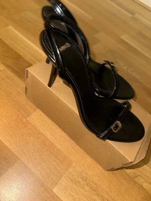 Svarta pumps med smala remmar från MNG - Stilrena svarta pumps från MNG med smala remmar och liten spänndetalj framtill. Skorna har öppen tå och hög klack, perfekt för en trendig look. Tillverkade i glansigt material som ger en elegant finish. Använda 1 gång. 9 cm klack nypris 429 kr säljer för 300 kronor.