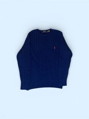 Mörkblå kabelstickad tröja Polo Ralph Lauren - Mörkblå kabelstickad tröja från Polo Ralph Lauren med rund halsringning och röd broderad logga på bröstet. Klassisk och tidlös design i mjukt material, perfekt för lager-på-lager-stil.