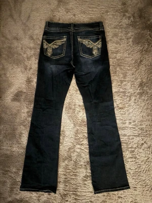 Mörkblå bootcut jeans med brodyr - Snygga mörkblå bootcut jeans med kontrastsömmar och broderade fickor bak med coolt vingmönster. Jeansen har låg midja och klassisk femficksdesign. Perfekta för dig som gillar en avslappnad men ändå trendig stil. Köpta för 360 kr💕💕 använda max 5 gånger