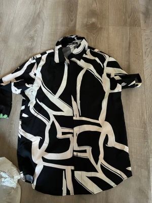 Svartvit skjorta från Manfinity - Säljer en svart skjorta från Manfinity med abstrakt vitt mönster. Skjortan har klassisk krage och knäppning framtill. Materialet känns slätt och lätt, perfekt för dig som gillar statement-plagg med unik design.