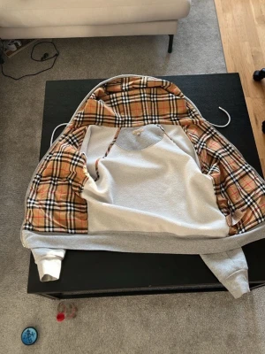 Grå hoodie med rutigt foder - Snygg grå hoodie med klassiskt Burberry-rutigt foder på insidan. Hoodien har huva med dragsko och långa ärmar. Utsidan är enkel och stilren medan insidan har det ikoniska beige, svarta och röda mönstret. Perfekt för dig som vill ha något unikt och trendigt.