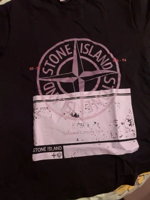 Svart Stone Island t-shirt med tryck - Snygg svart t-shirt från Stone Island med stort grafiskt tryck i vitt och lila på framsidan samt liten logga på bröstet. Klassisk passform och rund hals. Perfekt för dig som gillar streetwear och vill sticka ut med en ikonisk logga.