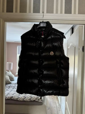 Moncler Väst Tibb (Size 2) - Snygg svart dunväst från Moncler med glansig finish och hög krage. Västen har dragkedja och knappar framtill samt två sidofickor med dragkedja. Klassisk Moncler-logga på bröstet. Perfekt för lager-på-lager och streetstyle.