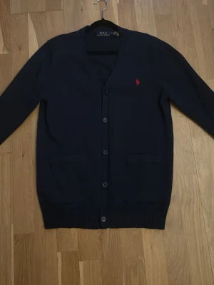 Mörkblå cardigan från Polo Ralph Lauren - Stilren mörkblå cardigan från Polo Ralph Lauren med knäppning framtill och två fickor. Klassisk röd logga broderad på bröstet. Långärmad modell i mjukt material, perfekt för lager på lager och en clean look.