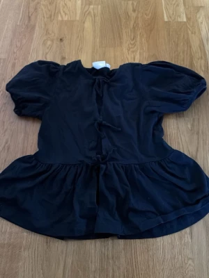 Svart blus med puffärmar och knyt - En svart blus med puffiga korta ärmar och knyt framtill. Blusen har en volang nedtill som ger en söt och trendig siluett. Perfekt för dig som gillar en avslappnad men ändå stylish look. Materialet känns mjukt och lätt, troligen bomull eller bomullsblandning.