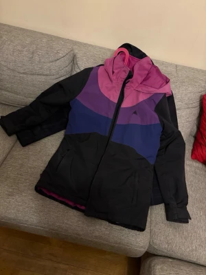 Färgstark skidjacka från Burton - Säljer en snygg vindjacka från Burton i svart, lila och rosa med dragkedja och huva. Jackan har två fickor framtill och är tillverkad i ett slitstarkt material som står emot vind. Perfekt för dig som vill ha en färgglad och sportig look.