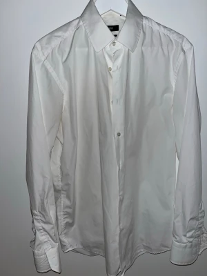 Vit skjorta från BOSS Regular Fit - Klassisk vit skjorta från BOSS i Regular Fit med knappar framtill och lång ärm. Skjortan har en stilren design med bred krage och är tillverkad i bomull, perfekt för en clean och snygg look.