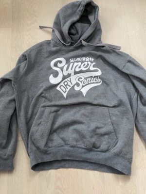 Grå hoodie Superdry  - Snygg grå hoodie från Superdry med stor vit logga och texten 'Superdry Sonics' på bröstet. Klassisk känguruficka framtill och justerbar huva med dragsko. Tillverkad i mjukt sweatshirtmaterial, perfekt för en avslappnad streetwear-look.M fits S