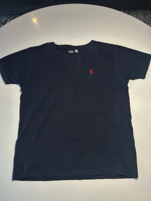 Mörkblå t-shirt från Polo Ralph Lauren - Klassisk mörkblå t-shirt från Polo Ralph Lauren med röd broderad logga på bröstet. T-shirten har rund halsringning och korta ärmar. Tillverkad i mjuk bomull för en skön känsla och enkel stil.