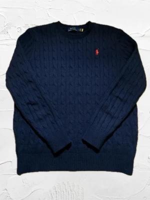 Mörkblå kabelstickad Polo Ralph Lauren - Mörkblå kabelstickad tröja från Polo Ralph Lauren med klassisk röd broderad logga på bröstet. Tröjan har rund halsringning, långa ärmar och ribbade muddar. Perfekt för dig som gillar stilrena och tidlösa plagg. Helt ny utan skador/missfärgningar. Några bilder i lite mörkare ljus och några i dagsljus. Snabb leverans 🚚📦⚡️.Vid köp av fler produkter = rabatt 💰📉✅.