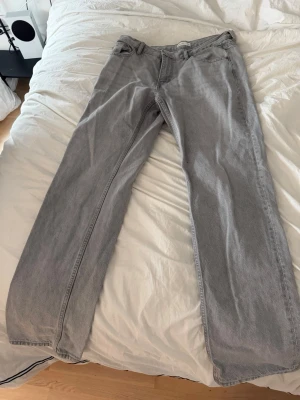 Grå raka jeans i klassisk modell  - Säljer ett par grå jeans med rak passform och fyra fickor. Jeansen har en klassisk look med knapp och dragkedja framtill samt bälteshällor. Materialet är denim och färgen är ljusgrå, perfekt för en chill och stilren vibe.