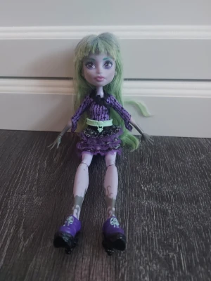 Monster High docka (Twyla) med lila outfit - Cool Monster High docka med långt ljusgrönt hår, lila och svart kläder med dödskallebälte och matchande lila skor. Dockan har detaljerade ben och armar med mönster och en rutig kjol med volanger.