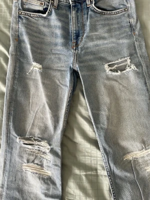 Ljusa slitna straight jeans H&M - Säljer ett par ljusblå straight high waist jeans från H&M med ankle-längd. Jeansen har flera slitningar och hål på benen för en trendig, avslappnad look. Tillverkade i denim med klassiska fem fickor och dragkedjegylf.