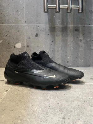 Nike Phantom Vision Elite fotbollsskor - Snygga svarta Nike Phantom Vision Elite fotbollsskor med silvriga detaljer och låg profil. Skorna har strumpliknande skaft, meshpaneler för ventilation och greppvänlig sula med orange dobbar. Perfekta för dig som vill ha kontroll och stil på planen.