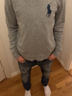 Grå sweatshirt från Ralph Lauren - Klassisk grå sweatshirt från Ralph Lauren med stor mörkblå logga på bröstet. Tröjan har rund halsringning, långa ärmar och ribbade muddar vid ärmslut och nederkant. Perfekt för en avslappnad och stilren look. Skriv för mer info