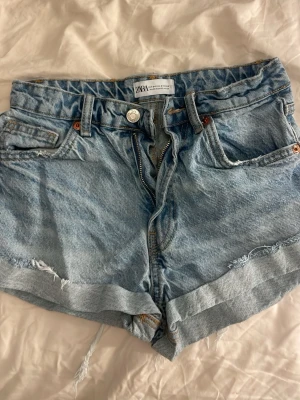 Ljusblå denimshorts från Zara - Säljer ett par ljusblå denimshorts från Zara med råa kanter och uppvikta benslut. Shortsen har klassisk femficksdesign, bälteshällor och dragkedja med knapp. Perfekta för sommaren och ger en avslappnad look.