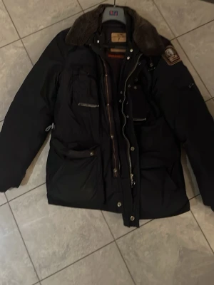 Svart dunjacka från Parajumpers - Säljer en svart dunjacka från Parajumpers med flera fickor, patch på ärmen och avtagbar pälskrage. Jackan har robust dragkedja, orange detaljer på insidan och quiltat foder. Perfekt för kalla vinterdagar och riktigt snygg streetstil.det är två delad 