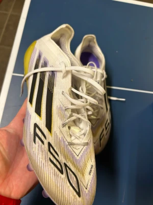 Adidas F50 Elite AG - Säljer ett par Adidas F50 fotbollsskor i vitt med svarta och gula detaljer samt lila inslag. Skorna har snörning och en smal, aerodynamisk form för snabbhet på planen. Ovandelen är i syntetmaterial och sulan har dobbar för bra grepp på gräs.