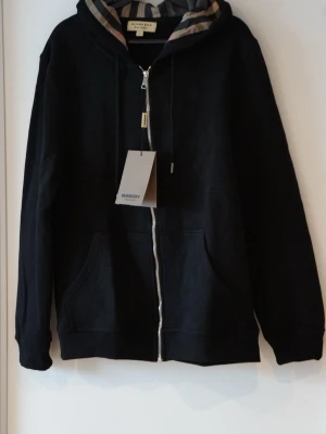 Svart zip hoodie från Burberry med dragkedja - Snygg svart hoodie från Burberry med dragkedja framtill och klassiskt rutigt foder i huvan. Tillverkad i mjukt bomullsmaterial och har två fickor framtill. Perfekt för dig som vill ha en stilren och exklusiv look med en touch av Burberrys ikoniska design. ❗️OBS❗️ - Skriv till mig innan köp! 🙌🏼